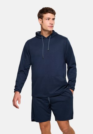 Joven con sudadera con capucha de manga larga azul marino y pantalones cortos a juego, de pie con una mano en el bolsillo, mirando hacia un lado.