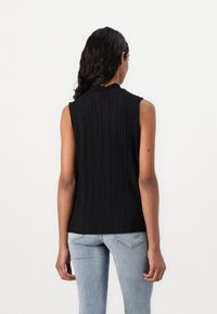 Haut sans manches noir avec des plis verticaux, un col montant et une texture douce. Porté avec un jean en denim bleu clair, mettant en valeur une silhouette ajustée.