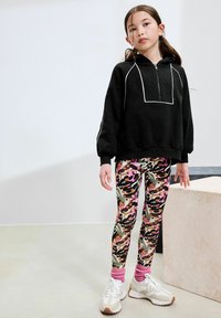 Svart zip-up hoodie med kontrasterande vitt sömnad, mönstrade leggings i rosa, gröna och bruna nyanser, samt vita sneakers med beige detaljer.