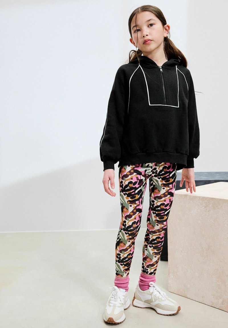 Fille aux longs cheveux portant un sweat à capuche noir, des leggings colorés à motif animalier, des chaussettes roses et des baskets blanches, debout à l'intérieur.