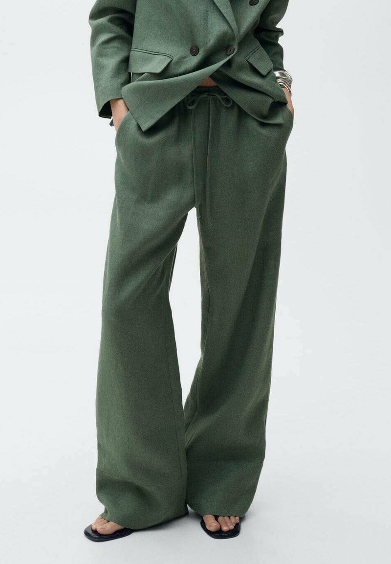 Personne portant un pantalon large vert olive et un blazer croisé assorti, les mains dans les poches et des sandales noires à bouts ouverts.