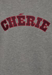 Grijze stof met een zachte textuur, voorzien van gedurfde, gebogen rode borduursels die "CHÉRIE" op de borst spellen.