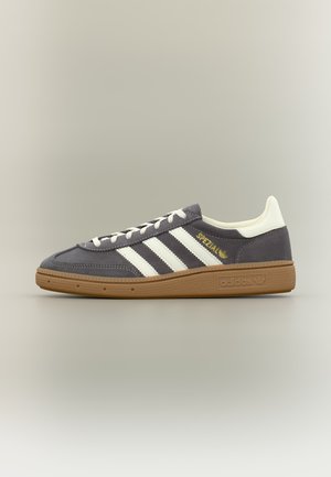 Sneaker Adidas Spezial grigia con strisce bianche, tallone bianco, scritta dorata “Spezial” e suola in gomma marrone, raffigurata su sfondo neutro.