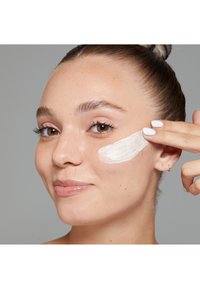 Creamy white product applied to cheek, smooth skin texture, natural makeup, light brown eyes, finger pointing to application area.