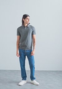 Grijze polo met korte mouwen, gedragen met blauwe jeans en witte sneakers. Model heeft lang haar en tatoeages, staand tegen een lichte achtergrond.