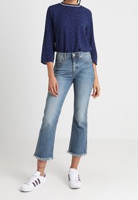 Blusa blu navy con pois rossi e rifiniture bianche, jeans croppati azzurri a vita alta con orlo sfilacciato, sneakers bianche con strisce scure.