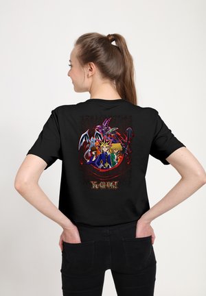 Giovane donna con coda di cavallo che indossa una maglietta nera Yu-Gi-Oh! con personaggi e draghi sul retro, mani posate nelle tasche posteriori.
