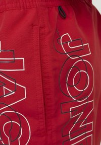 Pantalones cortos rojos con un bolsillo, presentando grandes gráficos de texto en blanco y negro. Tejido suave con un ligero brillo y una cinturilla elástica.