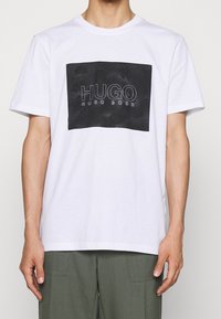 Bílý bavlněný tričko s černou obdélníkovou grafikou, která obsahuje text "HUGO HUGO BOSS", umístěná na texturovaném pozadí. Krátké rukávy.
