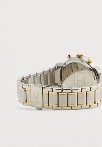 Rozsdamentes acél és arany színű fémből készült karóra, láncos karkötővel, zárható csattal és látható oldalsó gombokkal, világos háttér előtt.