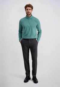 Pull sous-pull turquoise à zip quart et pantalon à motifs sombres, associé à des chaussures noires. Le pull présente des manches longues et une texture lisse.