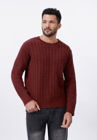 Kared maroon kampsun, millel on kabeli kootud muster, ümmargune kaelus ja pikad varrukad, koos tumesiniste teksadega valge tausta ees.