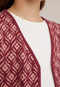 Bordeaux cardigan met een roze bloemenpatroon, open voorkant ontwerp en een gladde textuur, gedragen over een witte geribde top.
