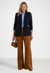 Donna dai capelli biondi che indossa un blazer blu navy su una camicia azzurro chiaro, pantaloni larghi marroni, scarpe marroni e tiene una piccola borsa marrone.