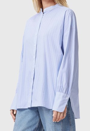 Femme portant une chemise ample à rayures fines bleu clair avec des poignets rayés contrastants et un jean bleu, debout devant un fond uni.