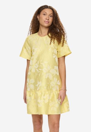 Kobieta o kręconych włosach, ubrana w żółtą sukienkę w kwiaty z krótkim rękawem i falbaną na dole, stojąca na jednolitym białym tle.