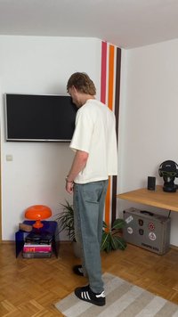 Um homem com uma camisa branca folgada e jeans azuis está ao lado de uma parede listrada colorida, uma lâmpada laranja e uma mesa de madeira com uma planta.