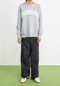 Felpa grigia con la scritta "GODDESS" in bianco e blu, abbinata a pantaloni larghi neri con motivo floreale e scarpe da ginnastica bianche.