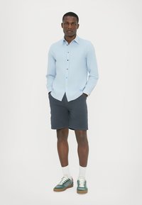 Ljusblå skjorta med knappar i flera färger, kombinerad med mörka shorts och gröna sneakers. Denna outfit har en avslappnad, casual stil.