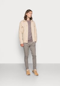 Chaqueta de algodón beige con dos bolsillos en el pecho, camisa de manga larga gris claro, pantalones gris ajustados, y zapatillas color marrón claro con detalles oscuros.