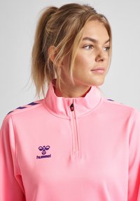 Felpa rosa con zip a quarti realizzata in materiale morbido, con logo viola e dettagli sulle spalle. Presenta un colletto alto e elementi di design a contrasto.