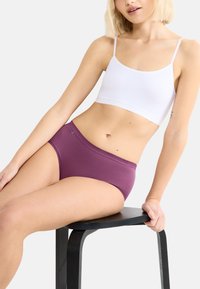 Fioletowe bawełniane majtki typu hipster w połączeniu z białym bawełnianym topem crop, obie części odzieży mają prosty, bezszwowy design. Modelka siedzi na czarnym stołku.