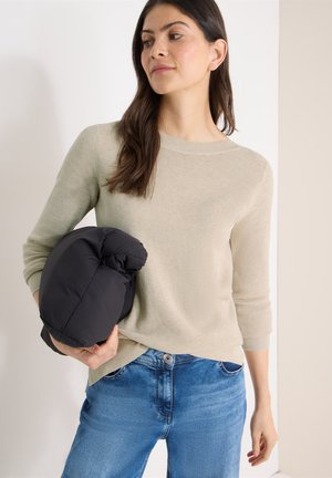 Femme en pull beige et jean bleu tenant une doudoune noire pliée, debout devant un fond neutre clair.