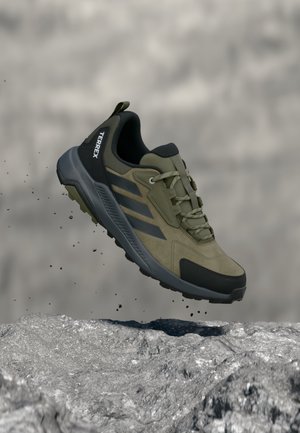 Chaussure de randonnée vert olive et noir avec marquage « TERREX », en l'air au-dessus d'un terrain rocheux avec de petites particules de terre tombant.