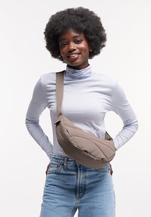 Taupe-gequiltete Crossbody-Tasche mit geschwungener Form, verstellbarem Riemen und Knopfdetaillierung, getragen über einem hellblauen langärmligen Oberteil.