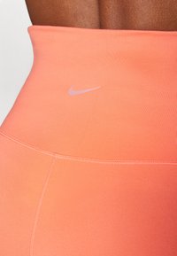 Høyt liv, ferskenfargede leggings med en glatt tekstur, som har en liten rosa Nike-logo og flate sømmer.