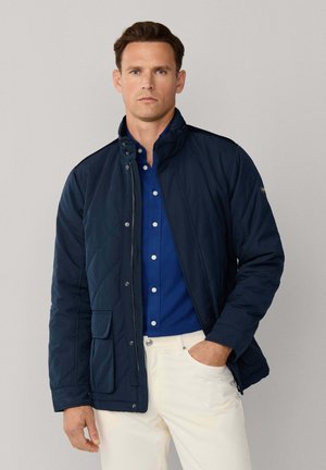 Donkerblauwe gewatteerde jas met een hoge kraag, een ritssluiting aan de voorkant en een drukknoopsluiting. Heeft een borstzakje en is gemaakt van zacht, lichtgewicht materiaal.