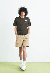 Mørkegrøn kortærmet t-shirt med en hvid broderet "N" og røde detaljer. Beige cargo shorts, hvide sportsstrømper og sneakers.