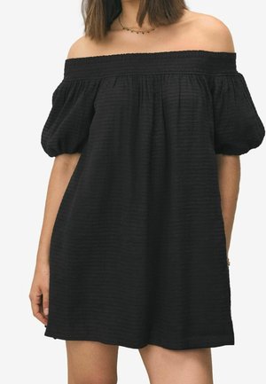 Femme portant une robe noire à épaules dénudées avec des manches courtes bouffantes et un tissu texturé, accompagnée d'un délicat collier en or.