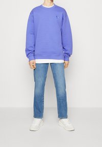 Felpa viola con polsini e orlo a coste, che presenta un piccolo logo di uccellino blu. Indossata sopra una maglietta bianca e abbinata a jeans azzurri e sneakers bianche.