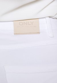 Jeans de denim blanco con una textura suave. Presenta una etiqueta rectangular etiquetada "ONLY" en un beige claro con letras en relieve y detalles de costura.