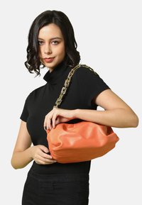 Borsa a tracolla in ecopelle arancione con un design morbido e soffice, dotata di una tracolla in catena dorata. La texture liscia e i dettagli cuciti sono ben visibili.