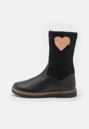 Bota negra de media pantorrilla con pie de cuero liso, superior de ante, parche en forma de corazón cosido en marrón y ribete marrón sobre una suela de goma negra.
