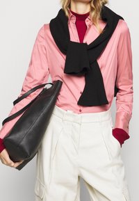 Sciarpa nera a coste appoggiata su una camicia rosa di raso con bottoni, abbinata a pantaloni larghi color crema. Viene tenuta una borsa tote in pelle nera strutturata.