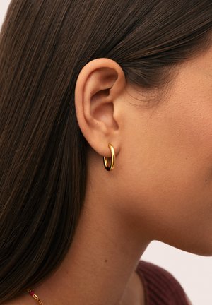 Primo piano dell'orecchio di una persona con capelli castani lisci che indossa un piccolo orecchino a cerchio d'oro e una maglietta bordeaux.