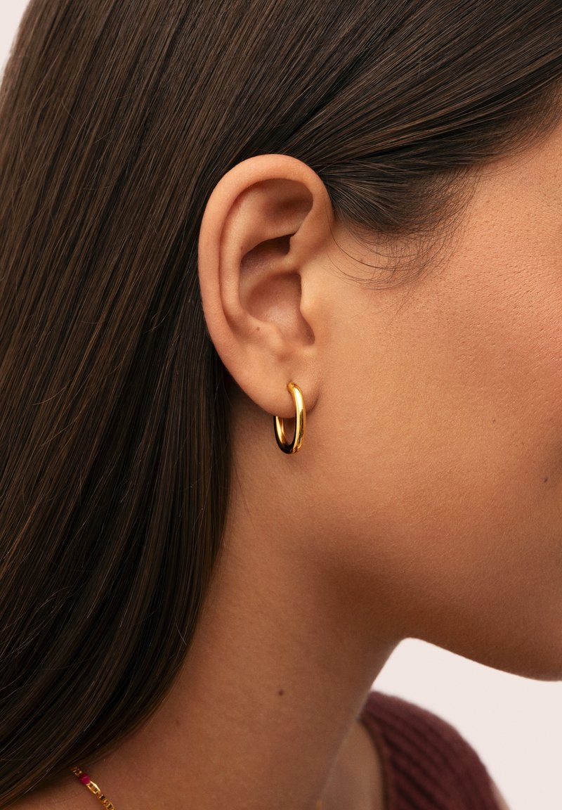 Primer plano de la oreja de una persona con cabello castaño liso que lleva un pequeño aro de oro y una camiseta color burdeos.