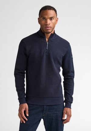 Mann steht frontal, trägt einen marineblauen strukturierten Viertel-Zip-Pullover und dunkelblaue Jeans vor einem einfachen weißen Hintergrund.