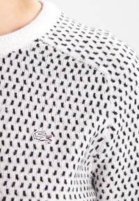 Vit stickad tröja med svarta prickar, med ribbat rundhals och en liten broderad logotyp på bröstet.