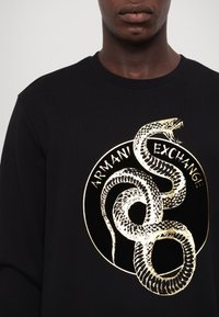 Närbild av svart sweatshirt med ett guldmetalliskt orm som är hopkrupen runt en guldcirkel och texten "ARMANI EXCHANGE" på bröstet.