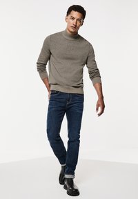 Pull en tricot gris avec un col montant, associé à un jean bleu foncé et des bottines noires à chevilles. Le look présente un design ajusté et un style décontracté.