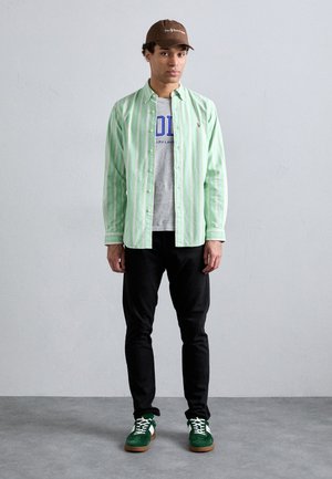 Camisa de botones a rayas verdes y blancas sobre una camiseta gráfica gris, pantalones negros y zapatos verdes con detalles blancos. Gorra marrón.