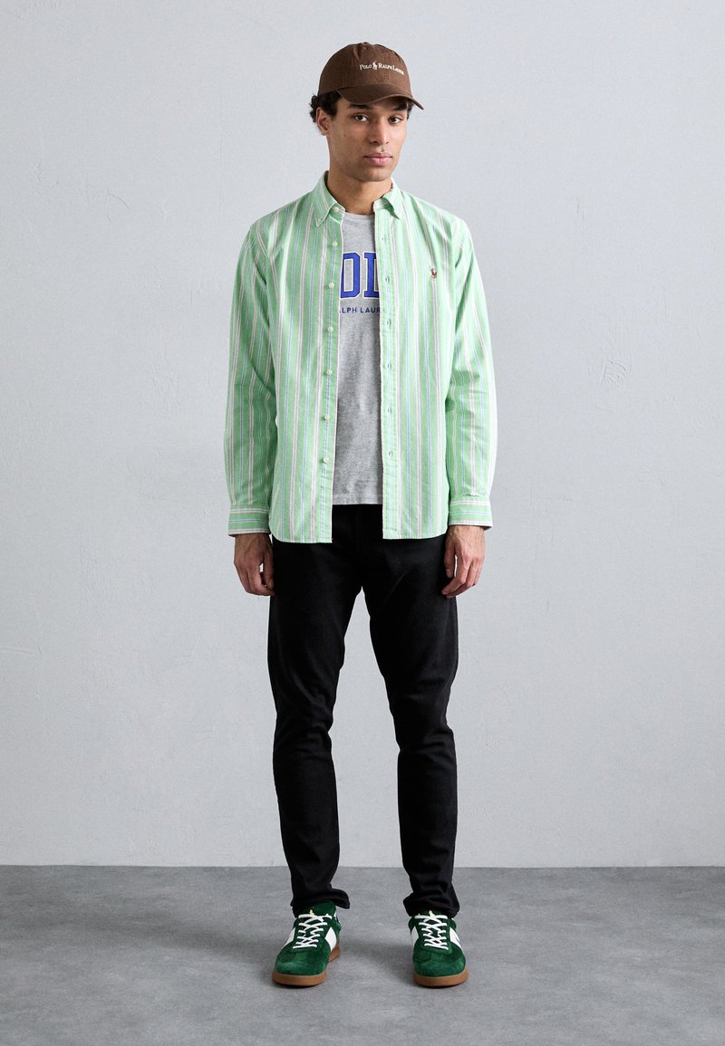 Camisa de botão às riscas verdes e brancas sobre uma t-shirt gráfica cinza, calças pretas e sapatilhas verdes com detalhes brancos. Boné castanho.