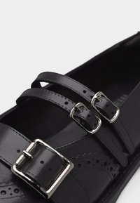 Chaussure en cuir noir avec trois boucles argentées et perforations décoratives, vue en gros plan sur un fond blanc.