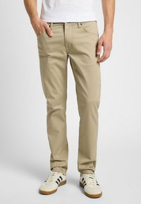 Chinos beige con un ajuste clásico, cinco bolsillos, cremallera frontal y sutiles detalles de costura. Llevados con zapatillas blancas y negras.