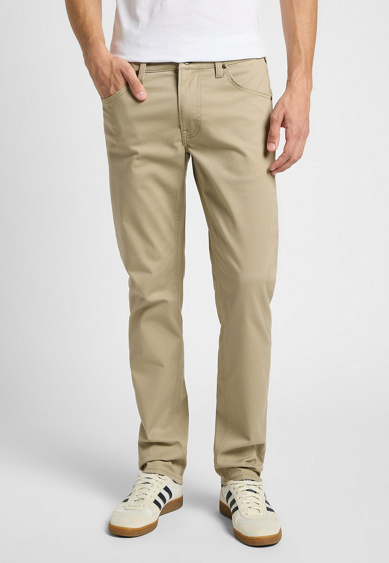 Chinos beige con un ajuste clásico, cinco bolsillos, cremallera frontal y sutiles detalles de costura. Llevados con zapatillas blancas y negras.