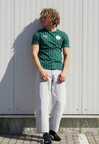Grünes kurzärmliges Puma-Shirt mit Rautenmuster, kombiniert mit hellgrauen weiten Hosen und schwarzen Sneakers; legere städtische Kleidung.
